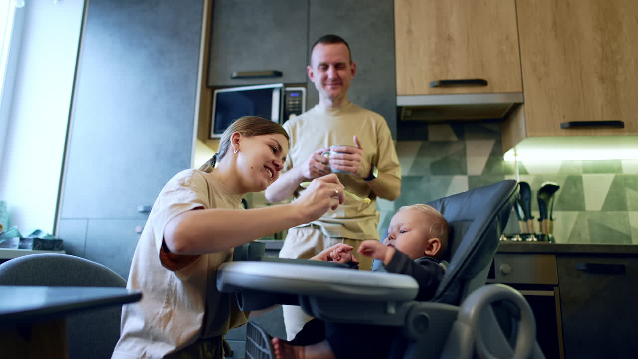 Feeding a little baby boy in the kitchen. Smiling mom gives the spoon to a kid and man stands behind with cup in hands.