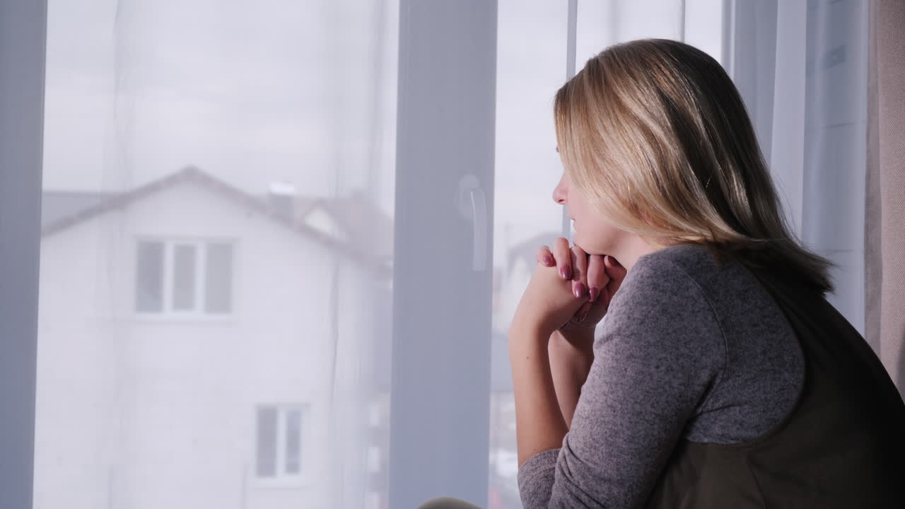 una mujer triste se sienta en el alféizar de la ventana mirando por la ventana a la casa 1