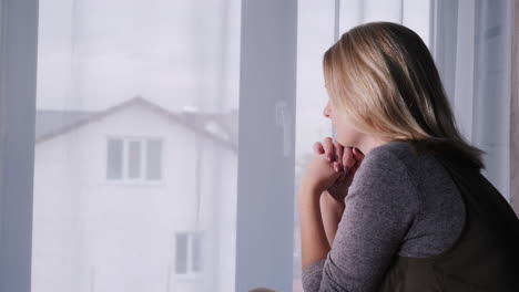 una mujer triste se sienta en el alféizar de la ventana mirando por la ventana a la casa 1