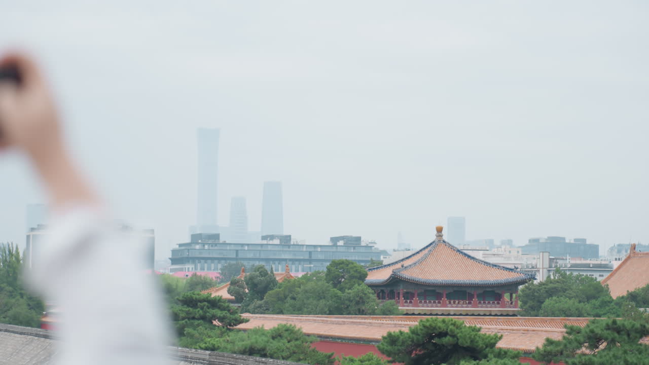Tetto di tempio, skyline distante. Turista che alza il telefono per fotografare la scena, torri moderne sfocate dietro una pagoda tradizionale, alberi verdi ed edifici bassi. Luce soffusa di cielo coperto, punto di osservazione panoramico.
