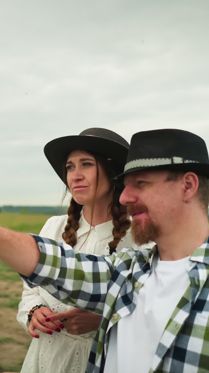 un cajón con una camisa a cuadros y un sombrero apunta con un pincel, apuntando. una mujer a su lado, con un vestido blanco y un sombreiro. se paran en un campo de hierba, con un lienzo parcialmente pintado en el fondo