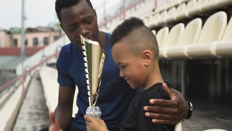 black trainer giving trophy to mixed race boy on stadium african american man giving cup and hugging mixed race boy while sitting on stadium after victory in match