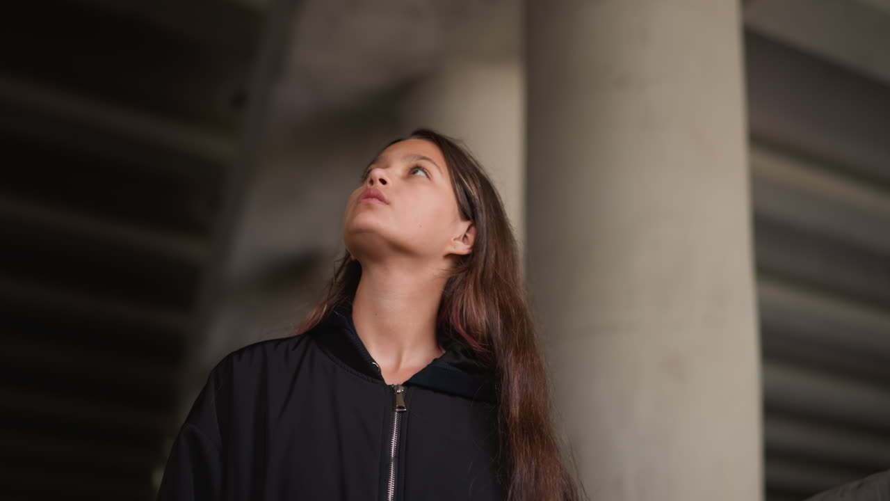 Pausa urbana chuvosa. Mulher observando o céu da ponte solenemente. Mulher contemplativa parada sob a ponte após a chuva. Cena urbana silenciosa com mulher olhando para cima em meio à humidade e iluminação suave