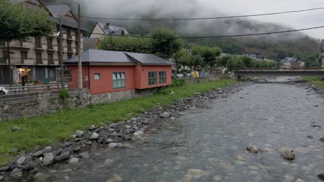 Die-Drohne-Konzentriert-Sich-Zunächst-Auf-Eine-Kleine-Hütte-Neben-Einem-Flussbett-Und-Steigt-Dann-Anmutig-Auf,-Um-Die-Charmante-Spanische-Stadt-Inmitten-Der-Malerischen-Pyrenäen-Zu-Enthüllen