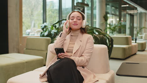 excited young businesswoman in headphones listening energetic music on smartphone in office lobby
