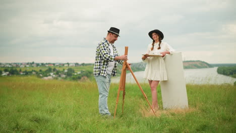 una pareja se prepara para pintar al aire libre en un campo verde exuberante con un. el hombre, con un sombrero negro y una camisa a cuadros, establece un caballete mientras que la mujer, con un vestido blanco y un sombreiro negro, sostiene una paleta de pintura