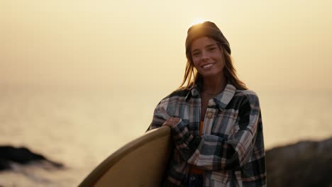 Portrait-of-a-blonde-girl-in-a-black-hat-and-plaid-shirt-who-holds-a-surfboard-stands-on-a-rocky-shore-near-the-sea-in-the-morning-and-looks-at-the-camera