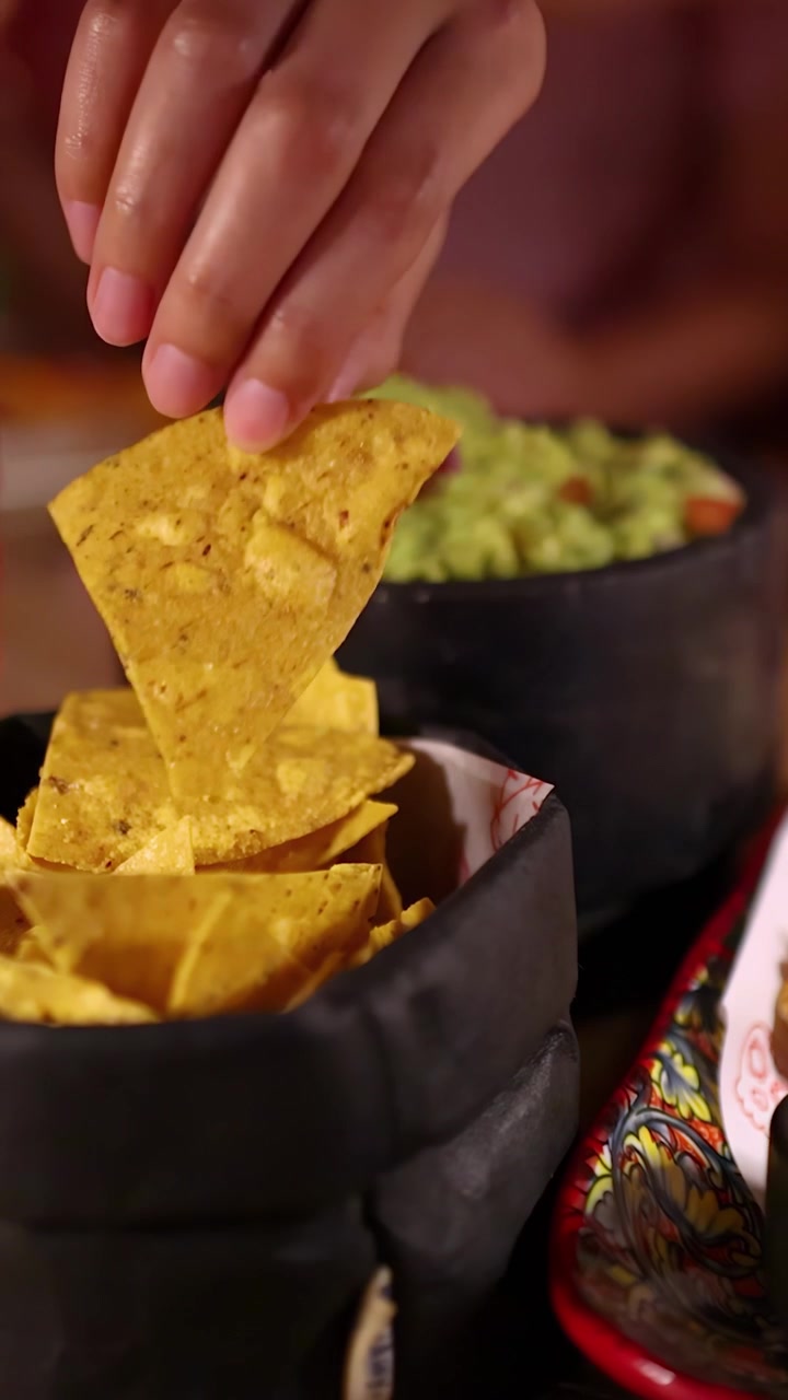 A hand reaches for a nacho chip, dips it into fresh guacamole in a rustic bowl, under warm, ambient lighting with a shallow depth of field
