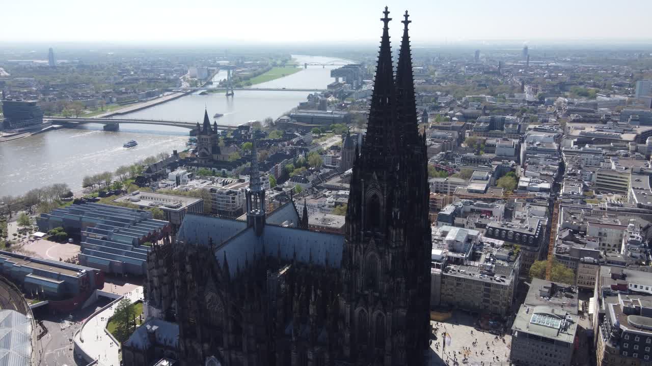 Aerial, flying close by gothic Twin towers of Cologne Cathedral - saint Peter Dom revealing view of Cologne riverfront cityscape, cargo barge and passenger ships moving on rhine river, Germany