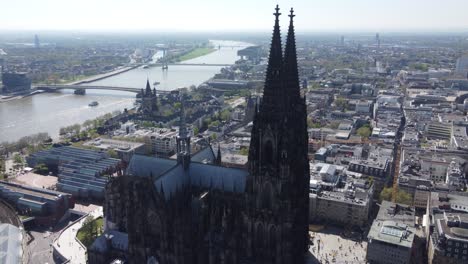 aerial, flying close by gothic twin towers of cologne cathedral - saint peter dom revealing view of cologne riverfront cityscape, cargo barge and passenger ships moving on rhine river, germany