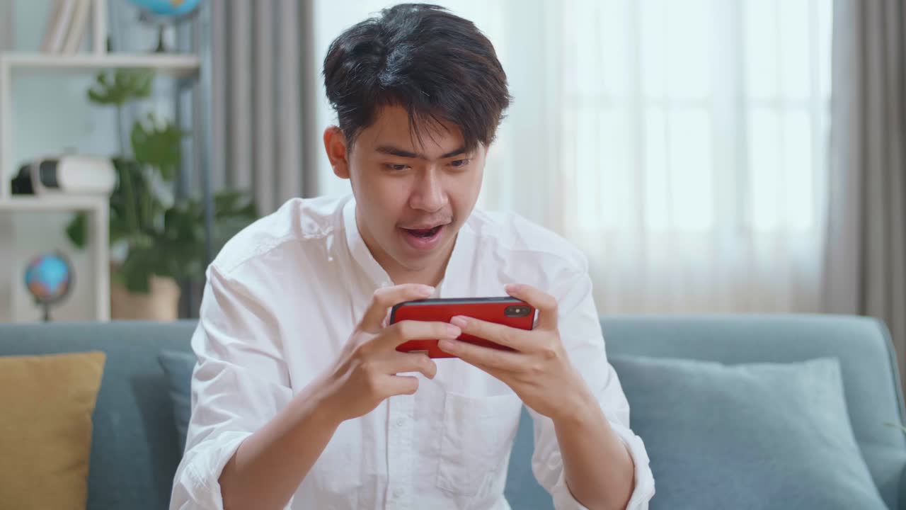Asian Man Celebrating Winning Game On Smartphone While Sitting On Sofa In The Living Room