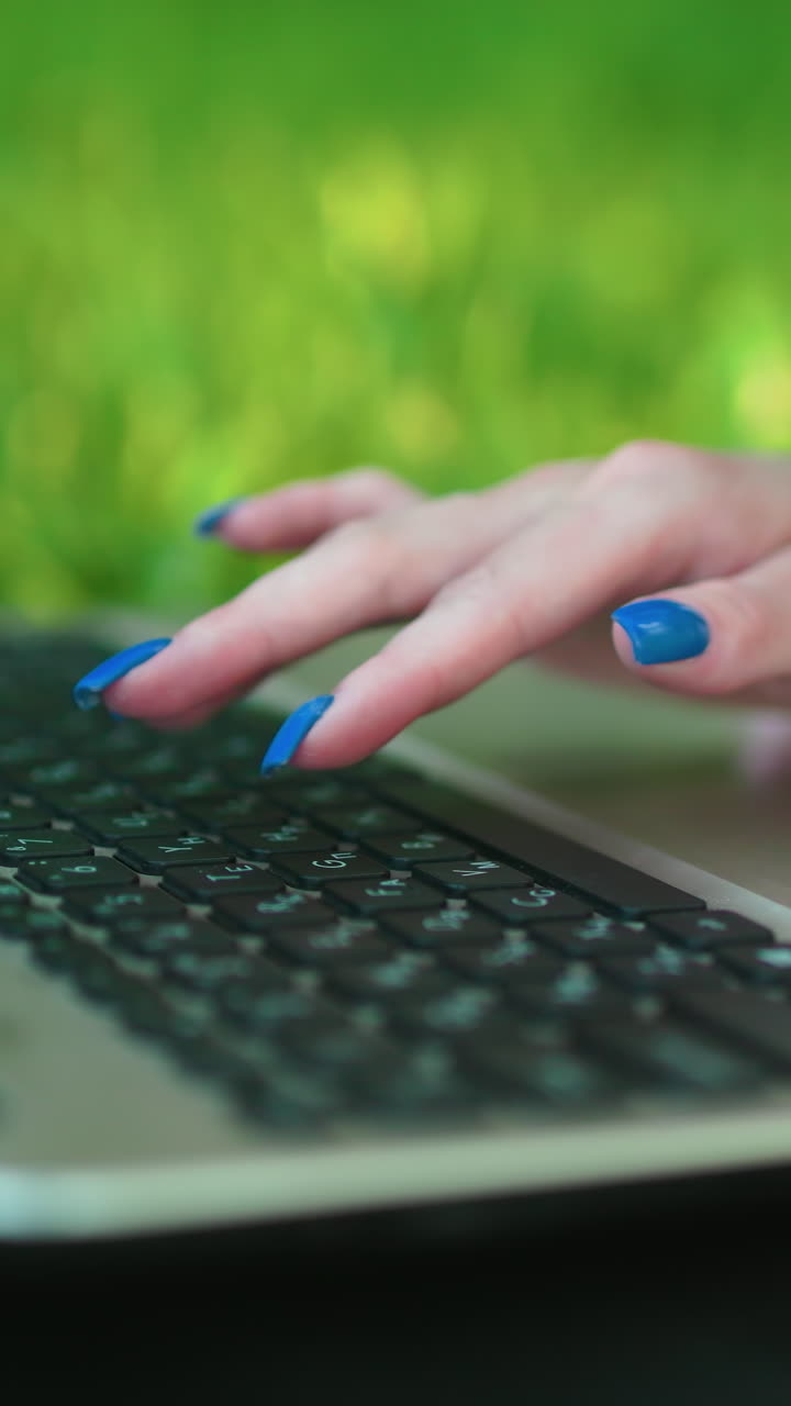 cerca de la mano de la señora con uñas pintadas de azul escribiendo en la computadora portátil mientras se acuesta en un campo cubierto de hierba al aire libre, creando una imagen de equilibrio entre la vida laboral y la vida y el trabajo remoto moderno con la naturaleza como fondo