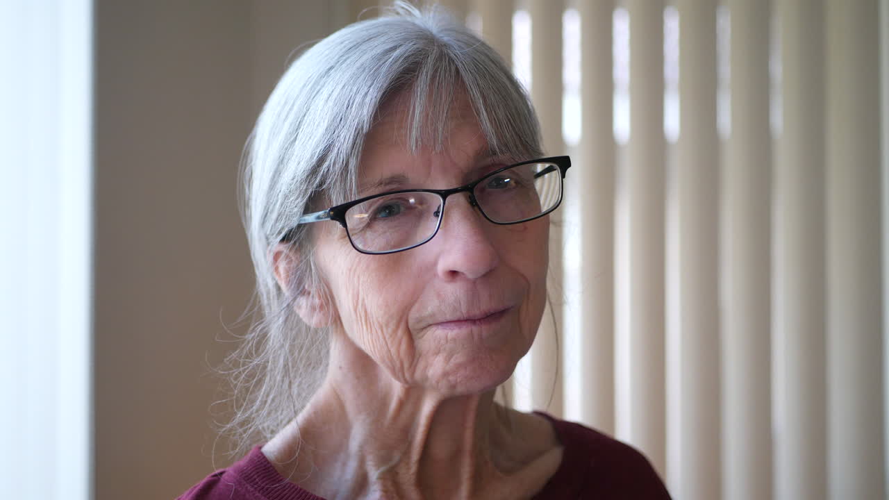 얼굴에 주름진 회색 머리와 안경을 쓴 백인 늙은 여자가 카메라를 바라보며 희망적이고 차분하며 행복하다