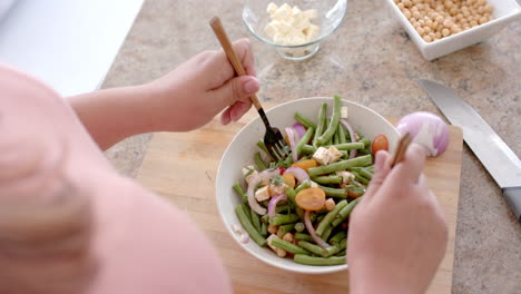 Midsection-of-plus-size-biracial-woman-tossing-bowl-of-feta-vegetable-salad-in-kitchen,-slow-motion