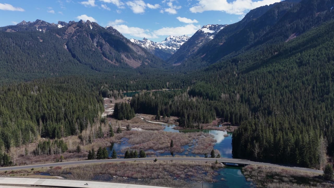 Ponte autostradale trafficato del Passo Snoqualmie sopra un fiume e una valle umida, fitta foresta sempreverde e cime rocciose innevate che si ergono ripide sotto un cielo blu. Washington, USA. Ampia ripresa aerea in ascesa