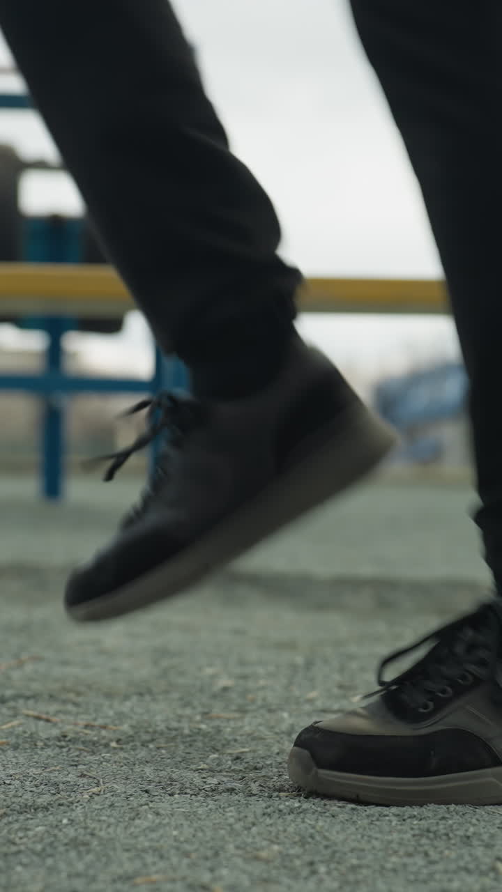 Close-up of a person's legs jogging rapidly, wearing black jeans and black sneakers, with benches around and distant houses visible in the background