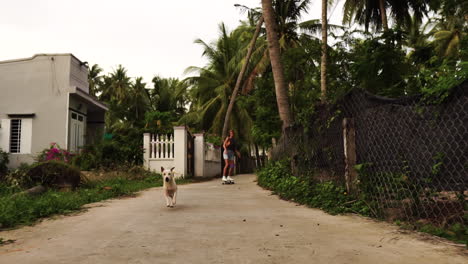 A-beautiful-young-caucasian-blonde-girl-is-riding-on-a-skateboard-following-by-a-dog-in-tropical-suburbs-in-South-East-Asia---low-angle-dolly-shot