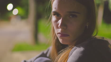 close-up of tired young woman gazing thoughtfully with a soft blurred background, illuminated by warm park lighting, capturing a moment of reflection and weariness in an outdoor evening setting