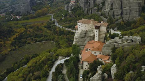 Aerial-view-around-the-Roussanou-monastery,-overlooking-winding-roads-and-foliage-forest-in-cloudy-Meteora,-Greece---high-angle,-circling,-drone-shot