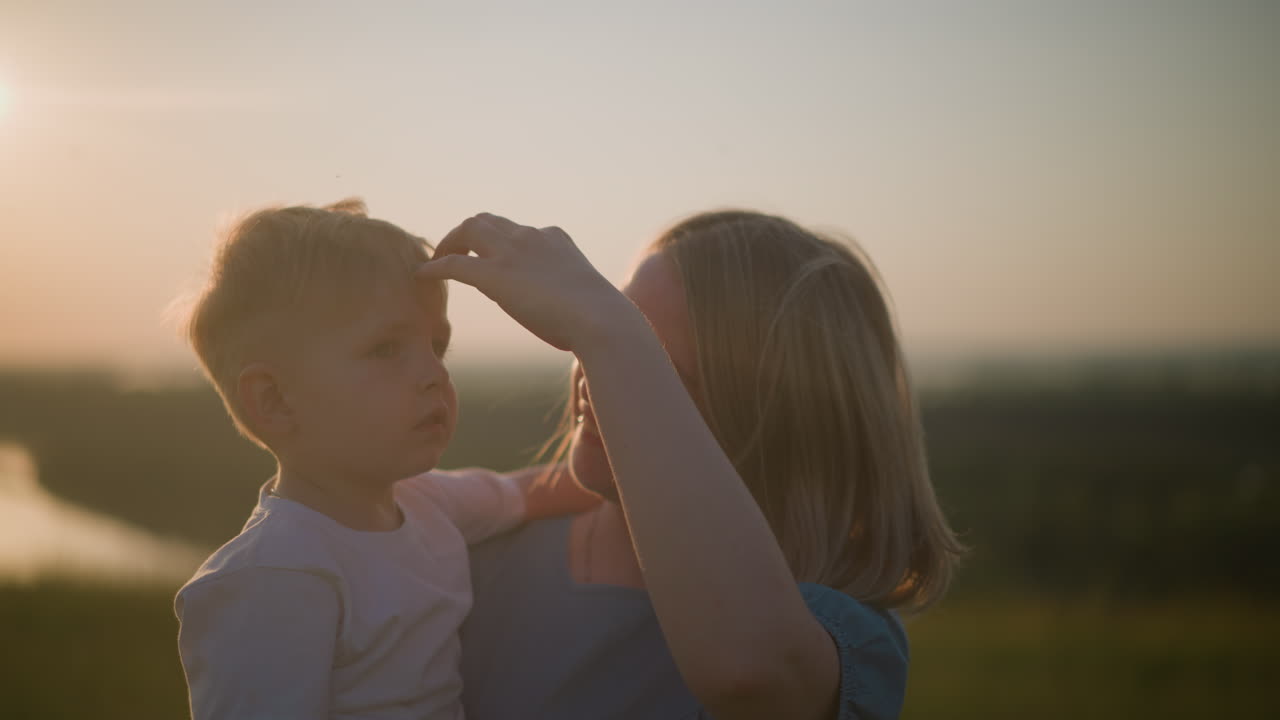 A mother and her young son in a grassy field at sunset. The woman, dressed in a blue gown, lovingly holds her little boy in a white shirt. She smiles warmly at him, while the boy gazes off,not smiling