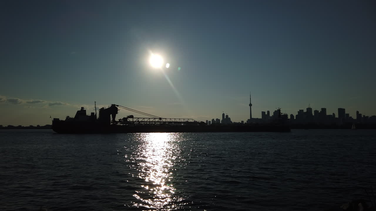 Long Great Lakes container ship preparing to enter Toronto harbour, silhouetted against the sun and Toronto skyline, static wide shot