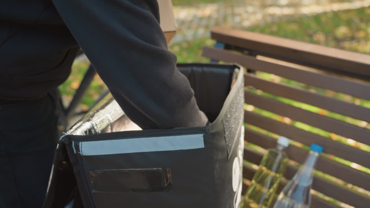 Corriere che preleva generi alimentari da una borsa termica su una panchina del parco, mette una scatola di cartone e bevande in bottiglia nel rivestimento termico, indossa una felpa scura con cappuccio e guanti con smartwatch visibile, ritiro contactless