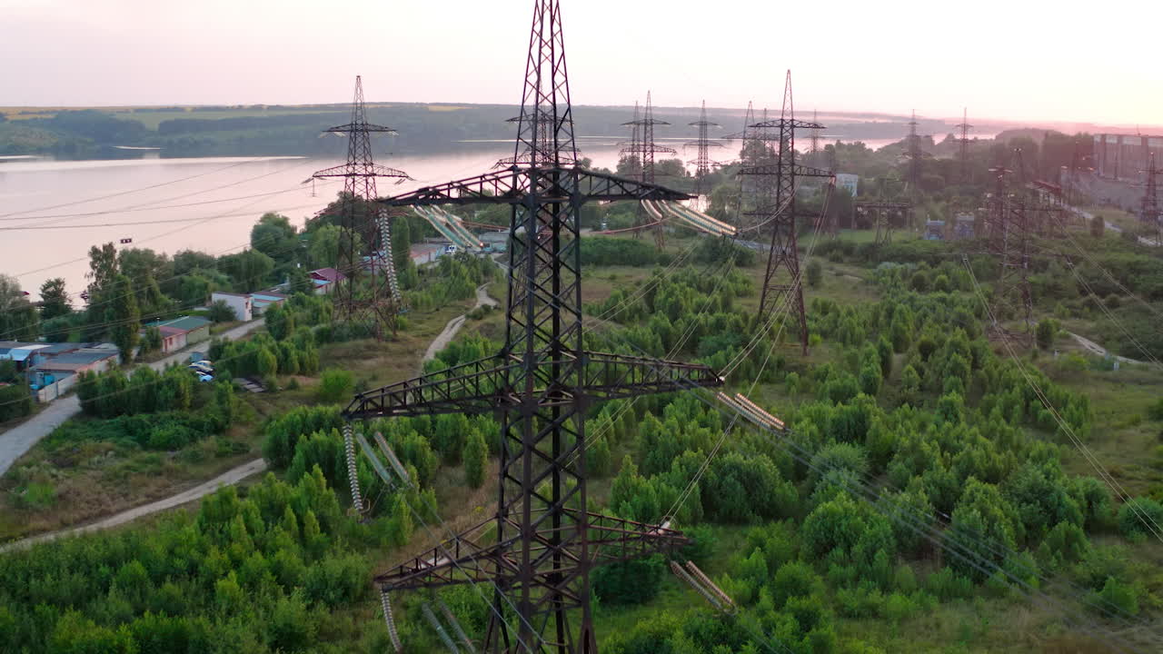 Structure of high-voltage lines outside the city at sunset. Trees and river. Camera motion back. Aerial view