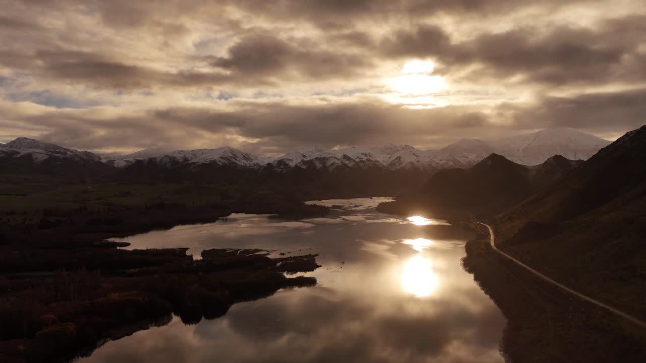 Waitaki River at sunset, calm water reflects cloudy sky, snow-capped mountains in background, New Zealand. Aerial drone