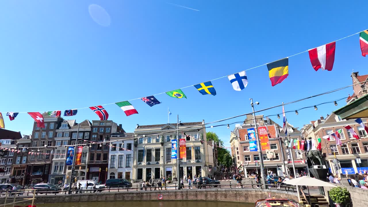 International flags hang above Amsterdam canal, historic buildings, bright daylight, static wide shot