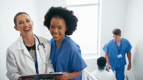 Portrait-Of-Female-Doctor-And-Nurse-With-Clipboard-Discussing-Patient-Notes-On-Stairs-In-Hospital