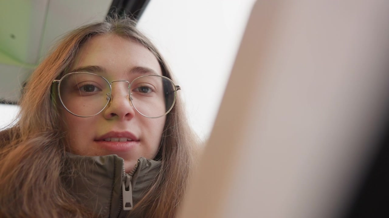 Jovem mulher branca em bonde olhando para o telefone, usando óculos e casaco de inverno com zíper em close-up de ângulo baixo com vibração de estudante espontânea, luz do dia suave através da janela, expressão sutil e trajeto autêntico