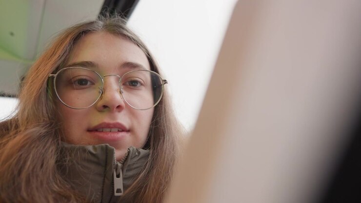 Giovane donna bianca sul tram che guarda il telefono, con occhiali e cappotto invernale con cerniera in un primo piano dal basso, atmosfera studentesca spontanea, luce naturale soffusa attraverso il finestrino, espressione sottile e autentico tragitto.