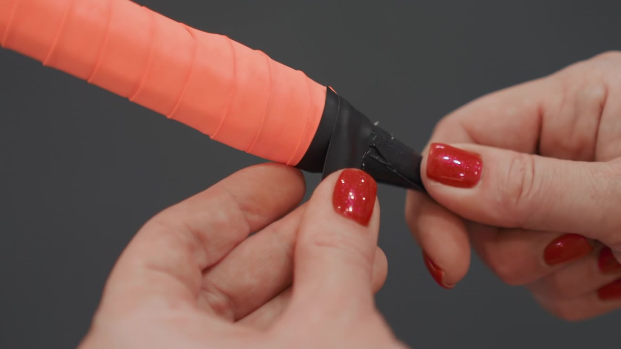 Primer plano de manos demostrando la aplicación de un overgrip de bádminton en el mango de una raqueta naranja, con sensación de demostración minorista y fondo de contraste oscuro, alineación precisa de la cinta, exhibición de producto para vendedores y entrenadores