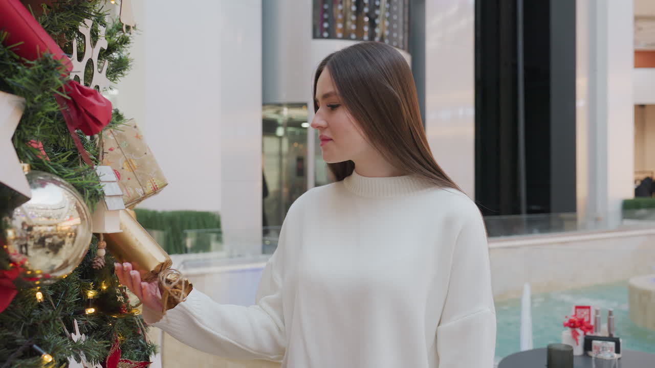 Young woman walks close to Christmas tree, gently holding golden decoration, with water springing and well-lit urban mall structures in background