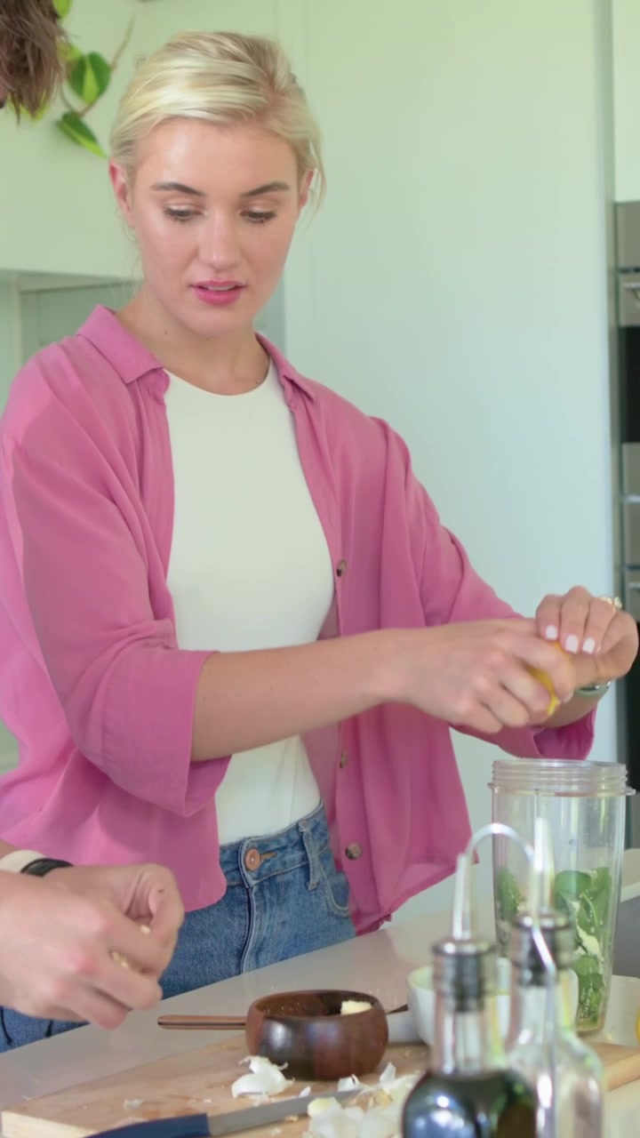 Video verticale: Donna che aggiunge verdure al frullatore e sprema un limone, mentre un adolescente trita l'aglio in cucina.