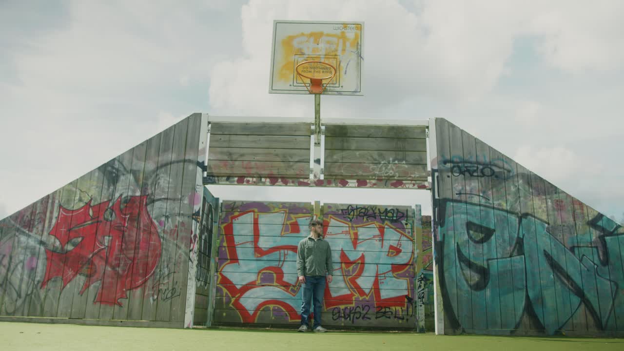 Front view of court with colorful graffiti walls, overcast daylight and visible basketball hoops, man stands at center posing