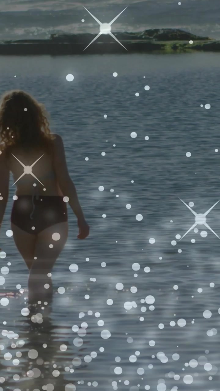 Donna adulta che parte dalla riva, addentrandosi nel mare poco profondo per il benessere, con scintille bokeh che la avvolgono.