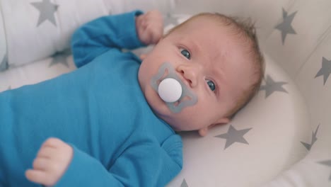 little child in blue bodysuit sucks dummy in cot with cocoon