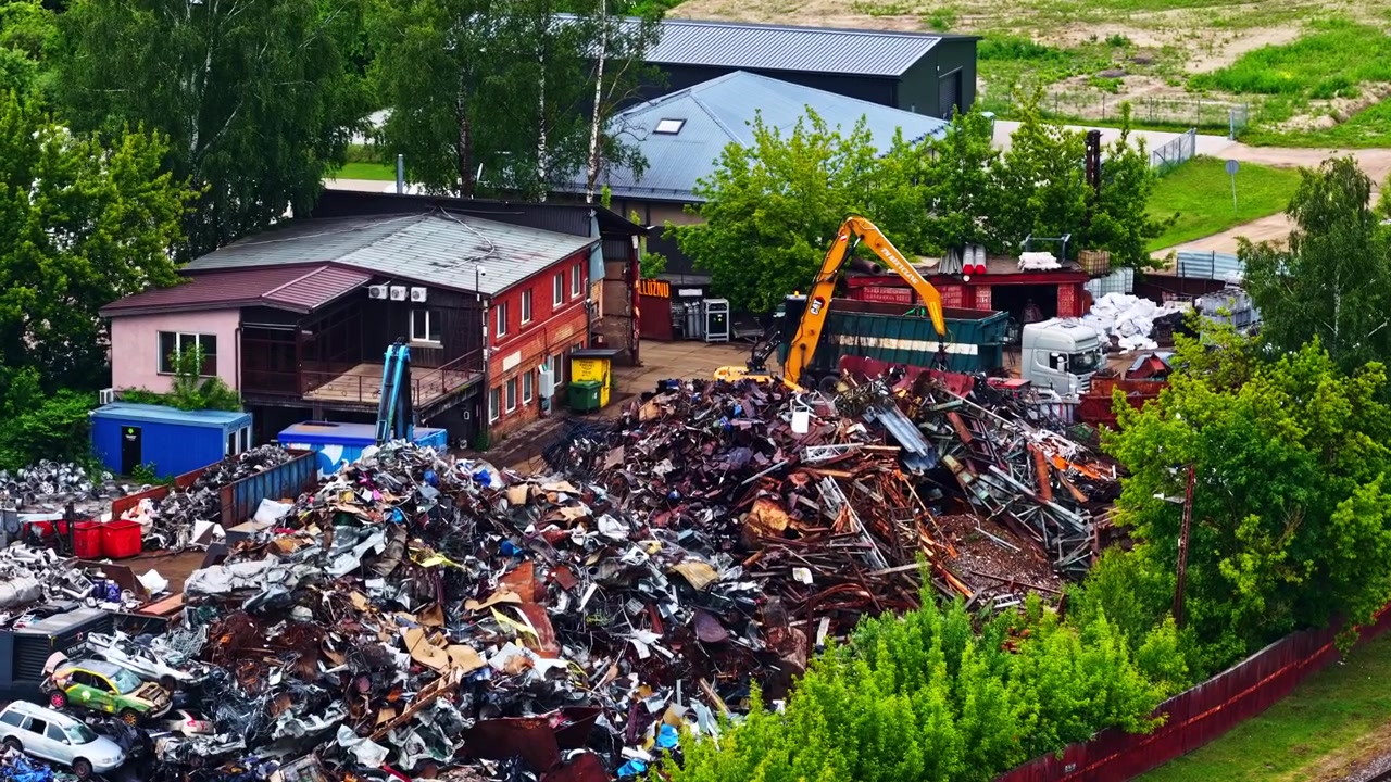 Un grande deposito di rottami pieno di cumuli di detriti metallici e veicoli vecchi che vengono lavorati da un escavatore. Ripresa aerea con drone