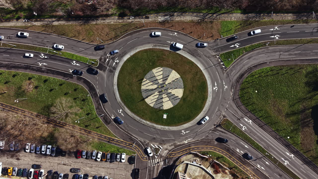 Cars move in circles around a roundabout with a patterned center and green grass in a city area during daylight hours