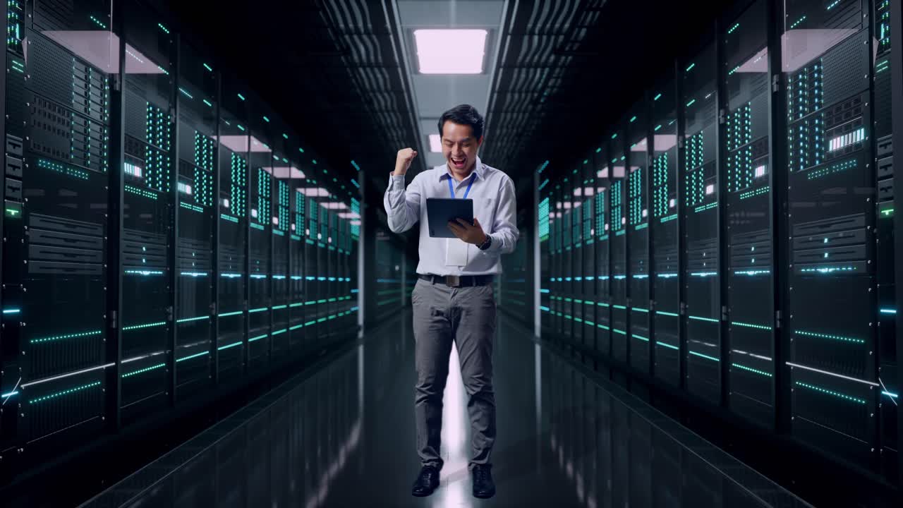 Helkropsbillede af en asiatisk mandlig professionel medarbejder, der står med sin tablet i et serverrum i et datacenter og hæver sin knyttede næve og råber "mål!", efter han har tastet på tabletten.