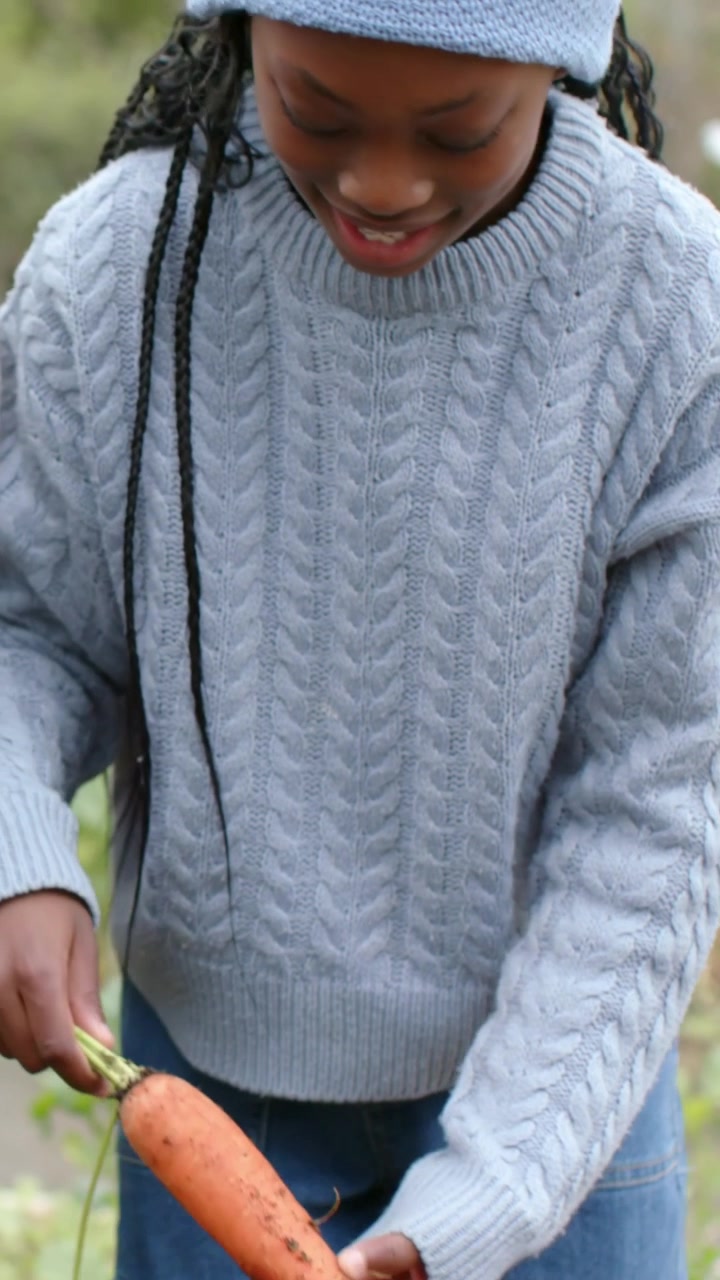 Vertikal video: Barn i sweater holder en trukket gulerod, tjekker, bøjer sig og plejer jorden i et højbed.