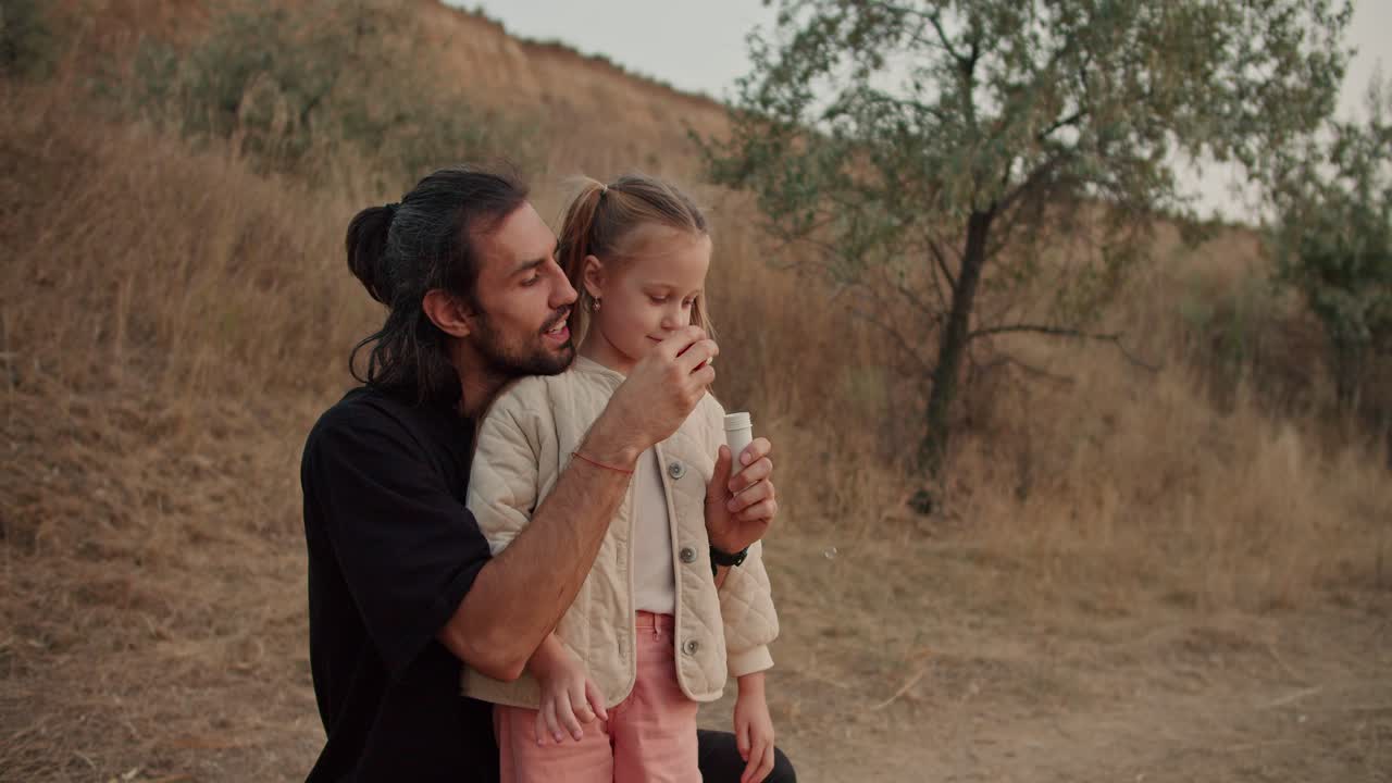A little brunette girl blows soap bubbles with her father during their picnic outside the city on a summer evening