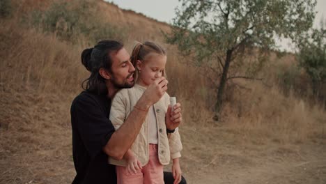 Una-Niña-Morena-Sopla-Pompas-De-Jabón-Con-Su-Padre-Durante-Su-Picnic-Fuera-De-La-Ciudad-En-Una-Tarde-De-Verano.