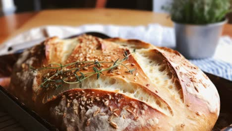 golden brown loaf of bread topped with fresh thyme and sesame seeds, resting in a dark baking pan on a wooden table, surrounded by a blurred plant backdrop