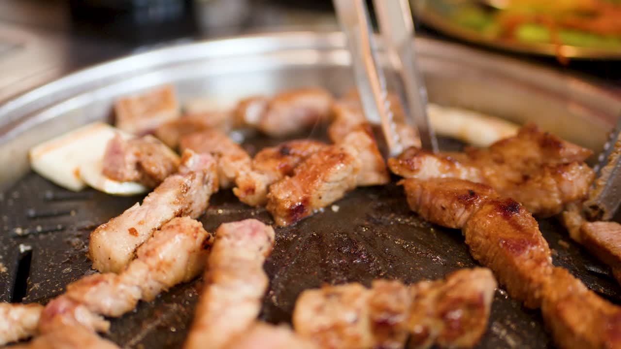 Pork belly pieces grilled on tabletop barbecue, tongs flipping meat, shallow depth, warm lighting