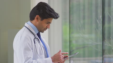 Male-Doctor-Wearing-White-Coat-Standing-In-Hospital-Corridor-Looking-At-Digital-Tablet