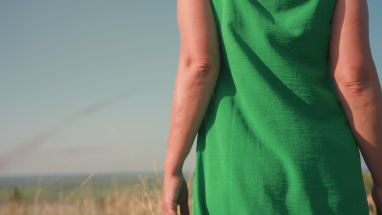 Primer plano trasero de una mujer con sombrero de sol blanco con cinta ajustando el tirante de un vestido verde mientras camina por hierba alta y seca bajo un cielo azul claro, con un suave desenfoque distante de un valle verde y un río abajo