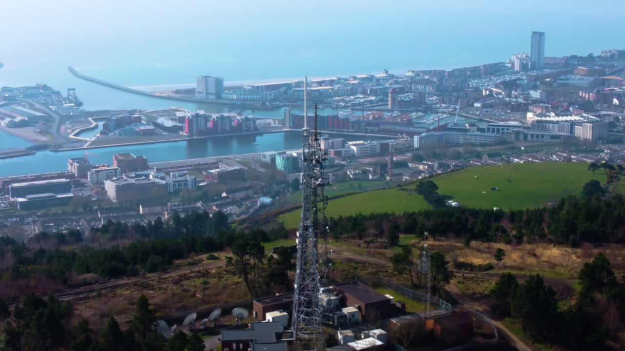 Sideways Aerial View of TV Radio Transmitter Tower High Above Swansea City with Marina in Distance. 5G Infrastructure for Smart Phones and Connectivity