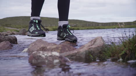 girl balances over wet stones to cross a stream with a hill in the background in scotland.